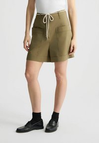 Pantaloni corti verde oliva con orlo ampio, due tasche frontali e un cordoncino. Abbinati a scarpe nere e calzini.