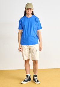 T-shirt en coton bleu, shorts cargo beiges avec poches latérales, baskets montantes noires, et casquette de baseball vert clair. Debout sur un sol jaune.