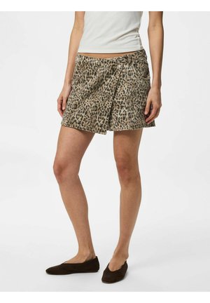 Femme portant une mini-jupe imprimé léopard, un top blanc sans manches et des chaussures plates noires, debout devant un fond uni.