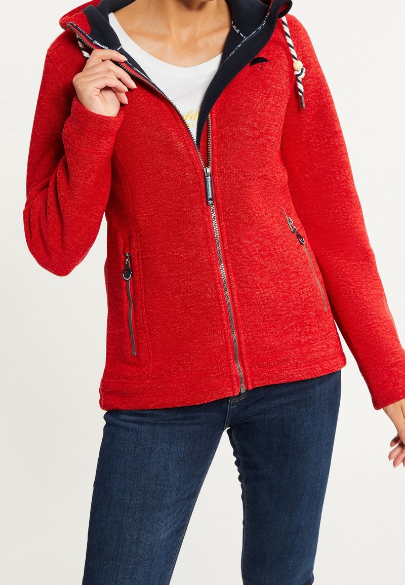 Femme portant une veste à zip rouge avec doublure bleu marine et un jean bleu foncé, tenant la fermeture éclair près du col contre un fond blanc.