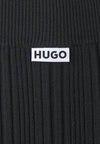 Czarny prążkowany materiał z białą prostokątną metką z napisem "HUGO" w pogrubionych czarnych literach. Tekstura dodaje wizualnego zainteresowania.