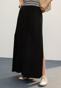 Crna maksi suknja s elastičnim pojasom, ravnog kroja s bočnim prorezima. Tkanina je glatka, lagana i lepršava.