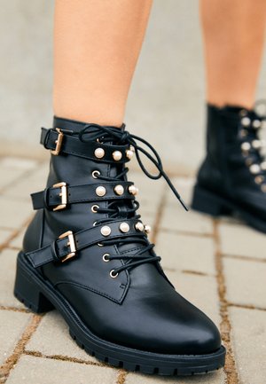 Bottines en cuir noir avec trois brides à boucle ornées de clous imitation perles, portées sur un trottoir pavé.