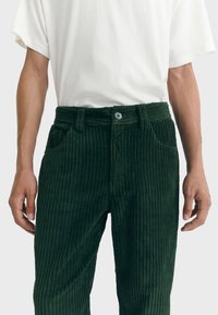 Pantalones de pana verdes con textura de cordones verticales, cierre de botón y trabillas para el cinturón. Los bolsillos frontales son visibles a ambos lados.