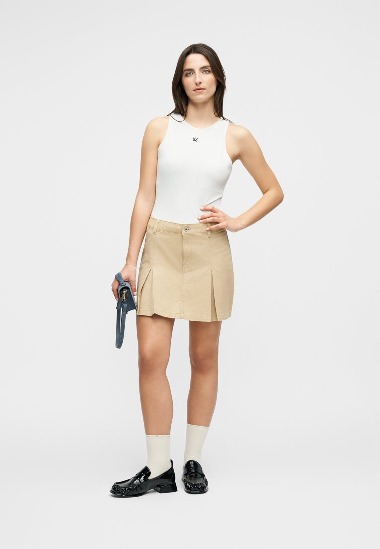 Femme debout, main sur la hanche, portant un haut blanc sans manches, une jupe plissée beige, des mocassins noirs avec des chaussettes blanches, tenant un petit sac à main bleu.