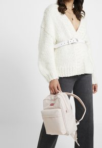 Beige reppu, joka on valmistettu kankaasta, etutasku ja kahva. Yhdistettynä valkoiseen paksuun neulepaitaan ja harmaisiin farkkuihin.