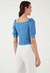 Blå floral bluse med puffærmer og kvadratisk udskæring, i et struktureret stof med hvidt blomstermønster. Stylet med hvide, løstsiddende bukser.