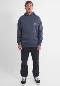 Sweat à capuche bleu marine en mélange de coton, doté d'une poche kangourou, de poignets côtelés et d'un petit motif brodé sur la poitrine. Associé à un pantalon foncé.
