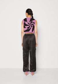 Femme aux cheveux tressés portant un haut court sans manches violet avec des motifs noirs en spirale, un pantalon cargo noir et des chaussures à talons roses, debout de dos.