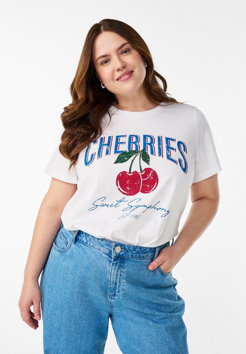 Pikkade pruunide juustega naine naeratab, kandes valget T-särki, millel on punased kirsid ja sinine tekst, ning kõrge vöökohaga siniseid teksaseid, käsi taskus.