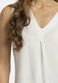 Top sin mangas blanco con escote en V y pliegue central, confeccionado en tela suave y con una textura sutil, que destaca un diseño simple.