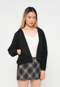 Crna zip-up hoodie sa bočnim džepovima, koja ima opušteni kroj i mekanu teksturu, u kombinaciji sa kariranim mini suknjom.