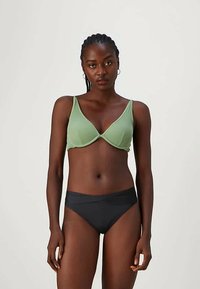 Ženska s kopletenimi lasmi, oblečena v svetlo zeleno modrček in črne hlačke, stoji pred enobarvno svetlo ozadje.