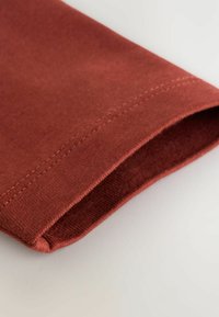 Nahaufnahme eines umgeschlagenen Saums aus rostfarbenem Stoff, der eine weiche Textur und eine saubere Naht entlang der Kante zeigt.