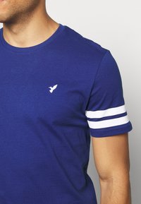 Modrá bavlněná tričko s malým bílým logem ptáka a bílými pruhovanými detaily na krátkých rukávech.