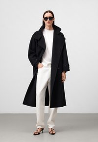 Schwarzer Wolls trenchcoat mit großem Kragen und Knopfdetails, getragen über einem weißen Hemd und einer cremefarbenen Hose, kombiniert mit schwarzgestriemten High Heels.