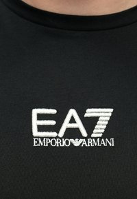 Must puuvillane t-särk, millel on valge tikitud logo tekstiga "EA7" ja "Emporio Armani", tekstuuriga õmblused ning ümmargune kaelus.