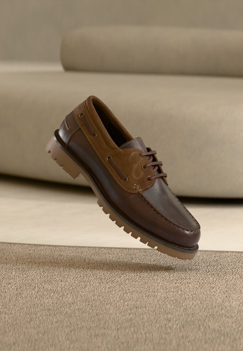 Καφέ δερμάτινο και σουέντ παπούτσι τύπου boat με κορδόνια, χοντρή ραβδωτή σόλα, τοποθετημένο πάνω από μπεζ χαλί και φόντο καναπέ.