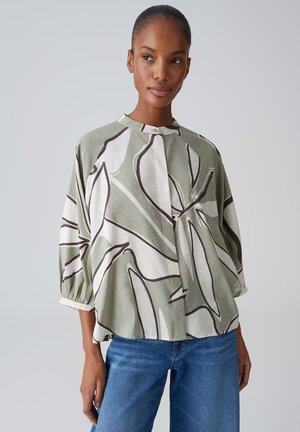 Donna che indossa una camicetta larga a maniche lunghe con motivo astratto di foglie in verde tenue, bianco e nero, abbinata a jeans blu.