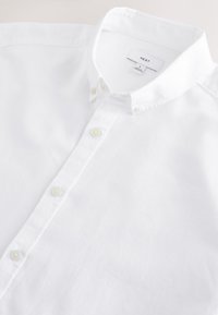 Chemise blanche à manches longues avec boutons, col boutonné et étiquette indiquant "NEXT", taille large, posée à plat sur une surface blanche.