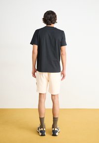 Black cotton t-shirt, short sleeves, round neckline, paired with cream-colored shorts. Model wearing gray and black sneakers with ribbed socks.