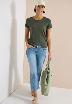 Femme portant un t-shirt vert olive, un jean bleu clair, une casquette beige, des chaussures marron clair, tenant un sac cabas matelassé vert, debout sur un fond beige et blanc.