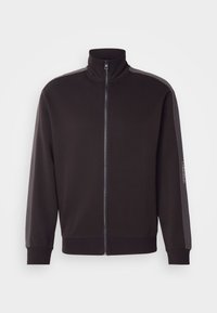 Veste noire zippée avec col montant et rayures gris foncé le long des manches, avec petit logo Calvin Klein sur le bras gauche.