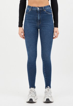 Woman wearing high-waisted blue skinny jeans, a black long-sleeve crop top, and white sneakers against a white background.