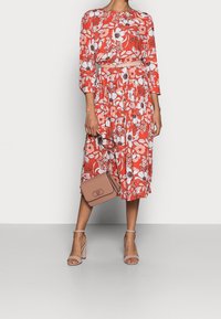 Robe florale rouge à manches longues, taille froncée et ourlet asymétrique. Accompagnée d'un sac bandoulière beige et de talons brides de cheville nude.