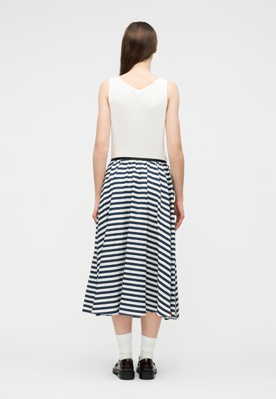 Γυναίκα με μακριά καστανά μαλλιά στέκεται πλάτη, φοράει λευκό αμάνικο τοπ, midi φούστα με μπλε ναυτικό και λευκές ρίγες, λευκές κάλτσες και μαύρα παπούτσια.