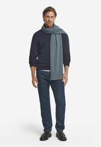 Marineblå sweater lagd med et blåt tørklæde,stående figur, mørkeblå jeans og sorte sko. Glatte teksturer med ribbede detaljer.