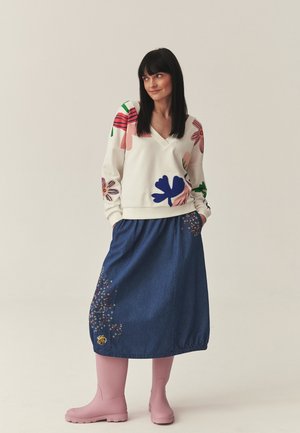 Kobieta w białej bluzie w kwiaty, dżinsowej spódnicy z haftem gwiazdy i pszczoły oraz różowych kaloszach, stojąca z rękami w kieszeniach.