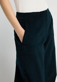 Sametové kalhoty v tmavě zelené barvě s jemnou texturou, vybavené bočními kapsami a bezešvým pasem s malým detailním knoflíkem.