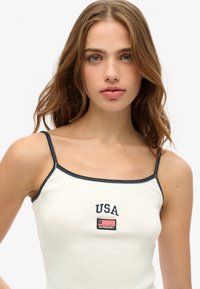 Weißes geripptes Tanktop mit navyblauem Nähten, das "USA" und ein kleines Flaggenlogo zeigt. Schmale Träger und taillierte Passform.