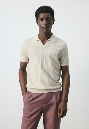 Jonge zwarte man staand, gekleed in een beige polo met korte mouwen met een structuur en mauve broek, tegen een effen lichte achtergrond.