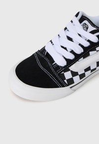 Sorte og hvite rutete sneakers har en overdel i semsket skinn og kanvas, hvite snøringer og en gummisåle med teksturert overflate.