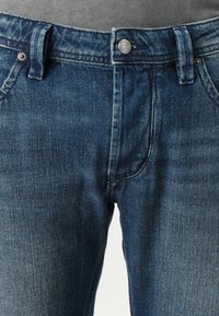 Tumedast sinine teksapüksid klassikalise viie-tasku disainiga, vöökohaga ja metallnupuga sulgemine. Kerge pleekimine nähtav.