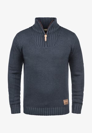 Mann trägt dunkelblauen gestrickten Pullover mit halbem Reißverschluss und schwarze Jeans, rechte Hand in der Tasche, linke Hand entspannt an der Seite.