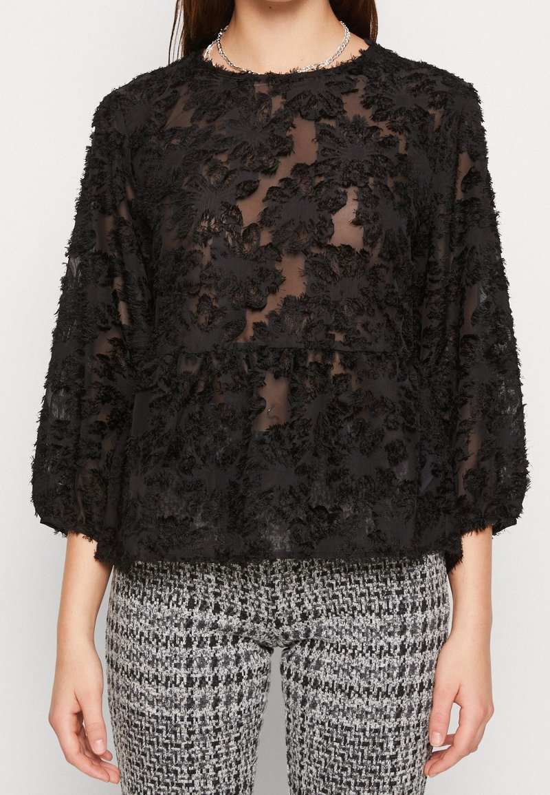 Femme portant un chemisier noir transparent à texture florale avec des manches trois-quarts et un pantalon à motif noir et blanc.