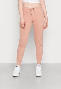 Person som bär ljusrosa Nike-sweatpants med vita Nike Air Max-sneakers som står på en grå golv.