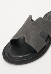 Grå stoff slide sandal med en bred kryssrem, åpen tå, og en svart gummisåle. Har teksturert materiale og minimalistisk design.