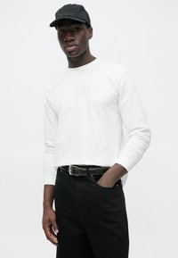 Ilgas baltas marškinėlis su įspaustu užrašu, derinamas su juodais džinsais ir juoda kepure. Apranga pasižymi lieknu siluetu ir lygia audinio tekstūra.