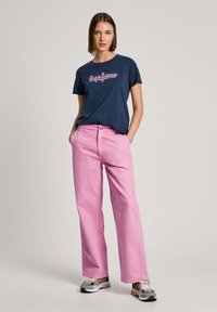 T-shirt azul-marinho com logotipo branco e rosa, combinada com calças largas de rosa claro e ténis multicoloridos com acentos pretos.