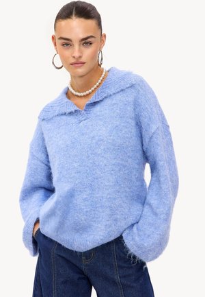 Pull oversized bleu clair en tissu doux et texturé. Doté d'un col et d'épaules tombantes. Associé à un pantalon en jean foncé.
