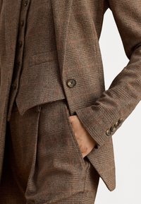 Ruskea herringbone-tyylinen bleiseri, jossa on istuva malli, rintatasku, ruskeat napit, jämäkät olkapäät ja sileä pinta.