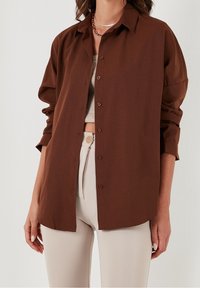 Camicetta marrone a bottoni realizzata in tessuto morbido, con una vestibilità rilassata, maniche lunghe e colletto classico, abbinata a pantaloni beige chiaro.