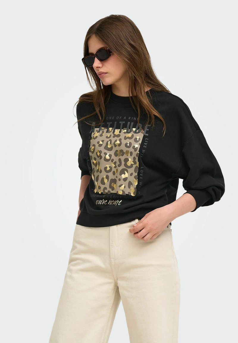 Femme portant un sweat-shirt noir avec imprimé léopard et texte, pantalon beige et lunettes de soleil noires, debout sur un fond uni.