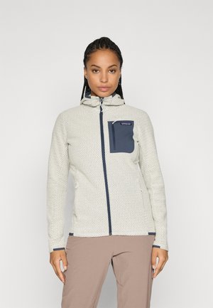 Frau trägt eine hellgraue gemusterte Zip-Jacke mit einer marineblauen Brusttasche und Kapuze, stehend vor einem schlichten Hintergrund.