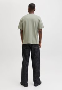 Mann mit kurzen Haaren, der ein hellgrünes kurzärmeliges Hemd, dunkle weite Jeans und schwarze Schuhe trägt, steht mit dem Rücken zum Betrachter vor einem schlichten Hintergrund.