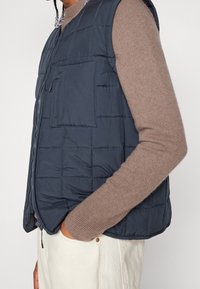 Marinblå puffväst med kviltad textur, framzip och flera fickor, bärs över en brun tröja. Har en slank, lätt design.